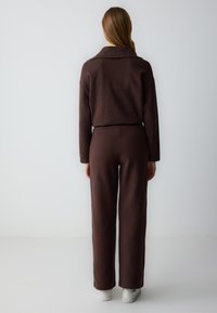 Salopette in maglia marrone con colletto, caratterizzata da una vestibilità ampia e gambe larghe. La texture è morbida, abbinata a sneakers bianche.