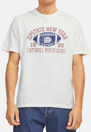 T-shirt en coton blanc avec un texte bleu et bourgogne et un graphique de football. Le texte comprend "New York de l'État," "1990," et "Montagnes Catskill."