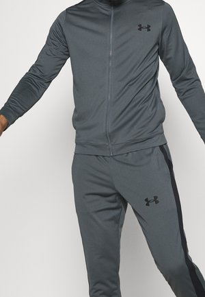Mężczyzna ubrany w ciemnoszary dres Under Armour z kurtką na pełny zamek i pasującymi spodniami z czarnymi paskami po bokach na jednolitym tle.