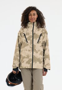 Beige camouflagejack met een hoge kraag, rits aan de voorkant en zwarte ritszakken; gladde textuur; gecombineerd met een zwarte helm.
