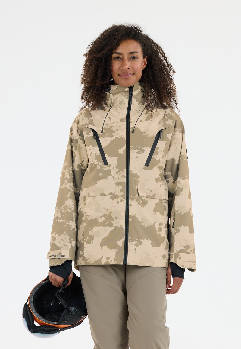 Beige camouflagejack met een hoge kraag, rits aan de voorkant en zwarte ritszakken; gladde textuur; gecombineerd met een zwarte helm.