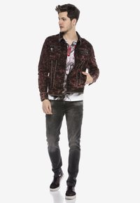 Rote Jeansjacke mit distressed Oberfläche, silbernen Hardware-Akzenten und sichtbaren Nähten. Getragen über einem grafischen T-Shirt und schwarzen Slim Jeans.