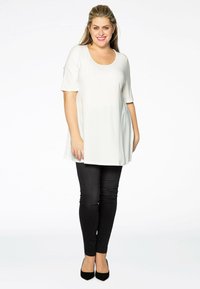 Weißes Shirt mit weitem Rundhalsausschnitt und kurzen Ärmeln, mit einem lässigen Schnitt. Kombiniert mit schmalen schwarzen Hosen und schwarzen spitz zulaufenden High Heels.