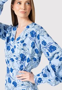 Blomstret blå kjole med V-udskæring, lange puffærmer og et let materiale med et mønster af mørkeblå blomster og grønne stængler.