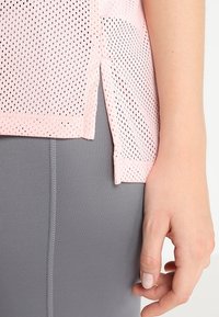 Top in rete rosa chiaro con piccole perforazioni, spacchi laterali e una texture morbida, abbinato a pantaloni grigi aderenti. Primo piano dei dettagli del capo.