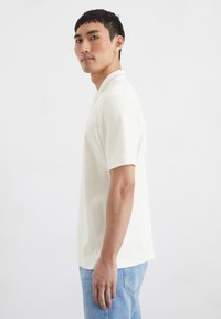 Helles beige Poloshirt mit kurzen Ärmeln aus glattem Stoff, mit klassischem Kragen und geradem Saum. Kombiniert mit hellblauen Jeans.
