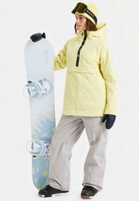 Vrouw in gele jas, beige sneeuwbroeken en zwarte handschoenen, houdend een witte en blauwe snowboard met bindingen, draag een gele muts en een skibril.