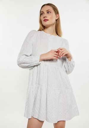 Młoda kobieta z długimi blond włosami, ubrana w luźną, jasną, długorękawową sukienkę w paski, stojąca na jednolitym białym tle.