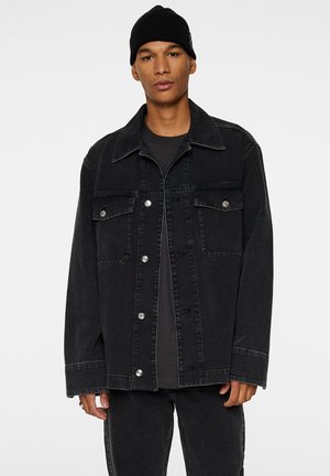 Black denim jacket with a relaxed fit, long sleeves, two chest pockets, silver buttons, and contrasting stitching. Worn over a black top.