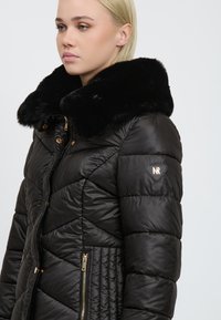 Quiltet sort pufferjakke med en stor sort faux fur-krave, guld hardware-detaljer og sidelommer med lynlås. Har et logo på ærmet.
