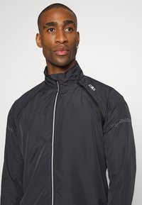 Schwarze leichte Sportjacke mit hohem Kragen, Reißverschluss, reflektierenden Akzenten und gepunktetem Muster auf den Schultern. Glatte Textur.