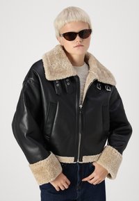 Veste courte en cuir noir avec un col et des poignets en fausse fourrure beige ; présente des accents à double boucle et une fermeture éclair.