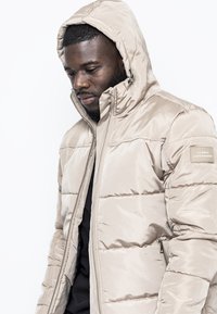 Giacca a soffietto beige con cappuccio, design trapuntato, zip frontale e tasche laterali. Presenta una toppa con logo sulla manica sinistra.