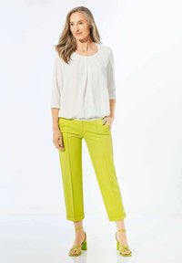 Weiße Bluse mit drei Viertelärmeln, kombiniert mit leuchtend grünen, taillierten Hosen mit glatter Textur und verkürzter Länge. Grüne Absatzsandalen runden den Look ab.