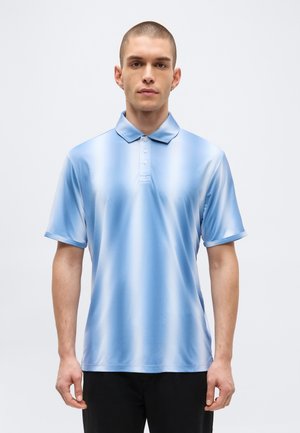 Jeune homme aux cheveux courts portant un polo rayé bleu clair et un pantalon noir, debout devant un fond clair uni.