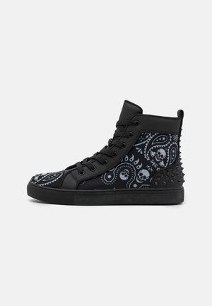 Schwarzer High-Top-Sneaker mit Paisley- und Totenkopfmuster, schwarzen Schnürsenkeln und schwarzen Spikes an der Ferse vor einem einfarbigen hellen Hintergrund.