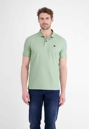 Mand med kort, krøllet hår iført en lysegrøn polo og mørkeblå jeans, stående mod en almindelig hvid baggrund.
