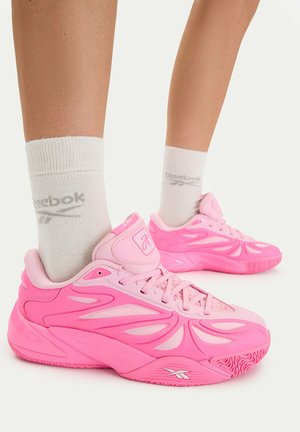 Nogi w białych skarpetkach Reebok i różowych sportowych butach Reebok z rzeźbionym wzorem i sznurowadłami na białym tle.