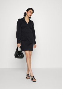 Vestido preto com colarinho, mangas bufantes longas e cintura ajustada. Usado com uma bolsa preta e sandálias pretas de tiras. Textura suave.