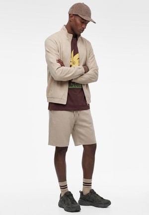 Man draagt beige jas, bijpassende korte broek, bruin grafisch T-shirt, beige pet, gestreepte sokken en zwarte sneakers, staand met gekruiste armen.