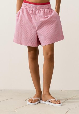 Personne portant un short rayé rose et blanc avec une ceinture rouge, debout pieds nus sur un sol carrelé clair.