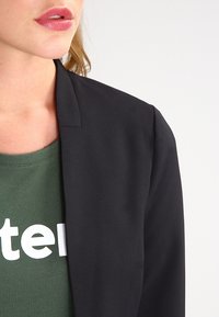 Femme portant un blazer noir sur une chemise verte avec un texte blanc partiellement visible, lèvres légèrement entrouvertes, fond clair.
