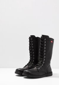 Botas negras de cuero hasta la rodilla con un acabado liso, cordones en la parte delantera, ojales plateados y cierre lateral con cremallera. Suela resistente con agarre texturizado.