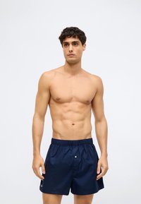 Μπλε ναυτικό μαγιό φτιαγμένο από ελαφρύ ύφασμα, με ελαστική μέση, ένα μπροστινό κουμπί και ένα μικρό λογότυπο στο πλάι.