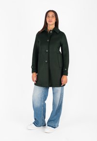 Cappotto lungo di colore verde scuro con colletto, cinque bottoni, due tasche laterali, dalla forma affusolata; abbinato a jeans larghi blu e sneakers bianche.