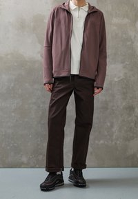 Brun zip-up jacka med huva, bärs över en vit skjorta med krage, kombineras med mörkbruna byxor och svarta sneakers. Slät tyg och enkel design.