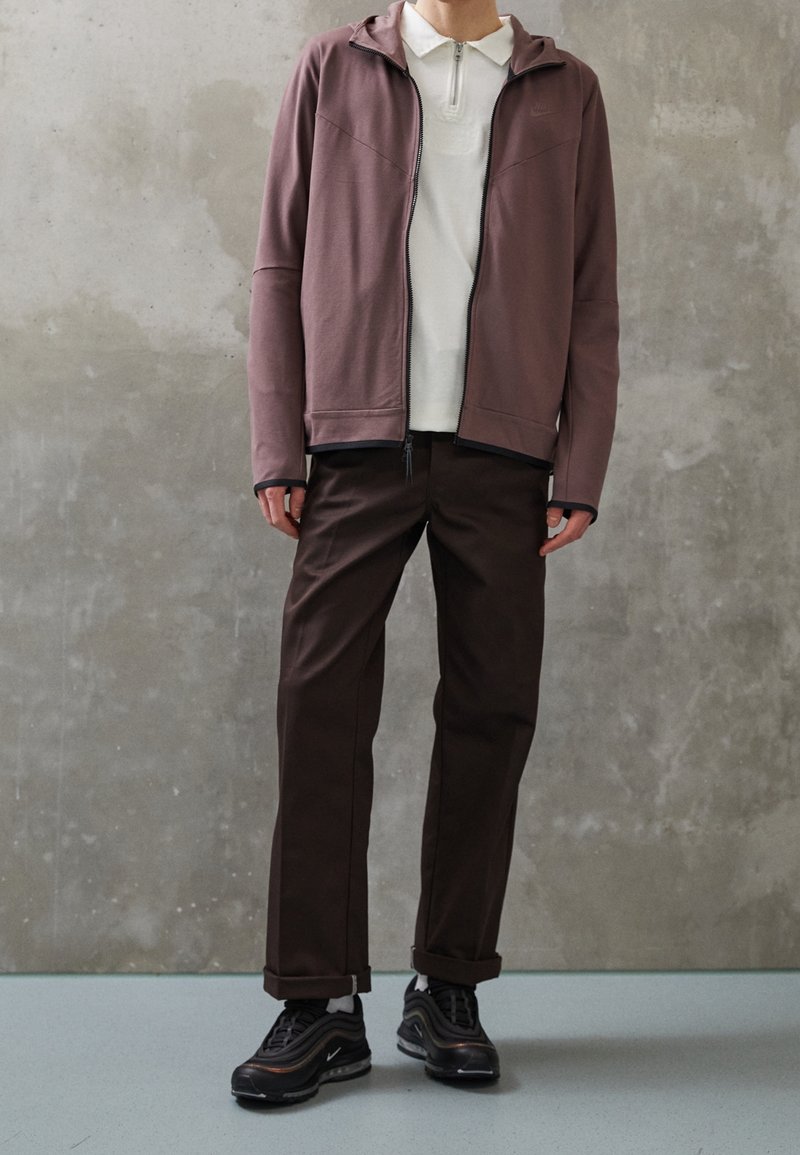 Brun zip-up jacka med huva, bärs över en vit skjorta med krage, kombineras med mörkbruna byxor och svarta sneakers. Slät tyg och enkel design.