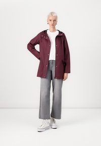 Lila vattentät jacka med huva, framdragkedja och tryckknappar, bärs över en vit skjorta och grå vida jeans. Vita sneakers ingår.