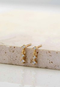 Goldene Ohrringe mit drei verbundenen Sektionen, von denen jede mit einem klaren, runden Edelstein besetzt ist. Glatte Textur des Goldes, reflektierende Oberfläche im Hintergrund.