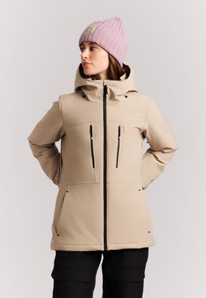 Femme portant une veste à capuche beige avec des fermetures éclair noires et un bonnet en tricot rose, regardant sur le côté avec les mains dans le dos.