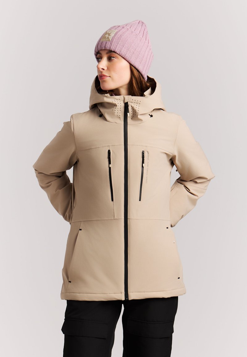 Woman wearing a beige hooded jacket with black zippers and a pink knit beanie, looking to the side with hands behind her back.