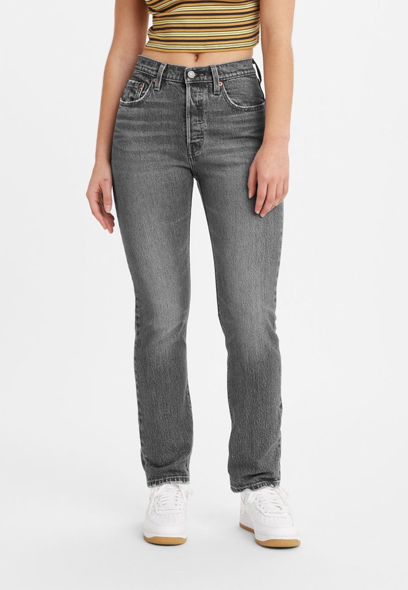 Femme portant un jean gris taille haute à jambes droites, un crop top rayé et des baskets blanches, se tenant devant un fond blanc uni.
