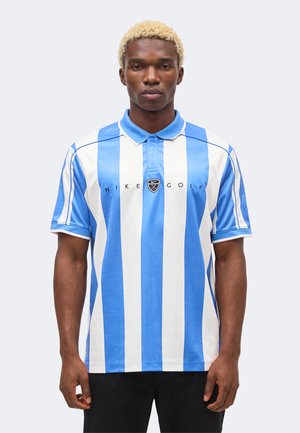 Homme aux cheveux blonds courts portant un polo Nike Golf bleu et blanc à rayures verticales, debout avec les bras détendus devant un fond uni.