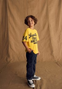 Geel katoenen T-shirt met marineblauw opdruk en nummer 84, gecombineerd met donkere cargobroek en zwarte sneakers met witte zolen. Eenvoudige achtergrond.