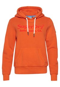 Sudadera naranja hecha de una mezcla de algodón, con un bolsillo tipo canguro, cordones y un gran logotipo de "SuperDry" en rojo en la parte delantera.