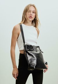 Schwarze Leder-Einkaufs-Tasche mit verstellbarem Riemen, glänzender Oberfläche, gerafftem oberen Teil und zwei seitlichen Schnallen zum Verschließen.