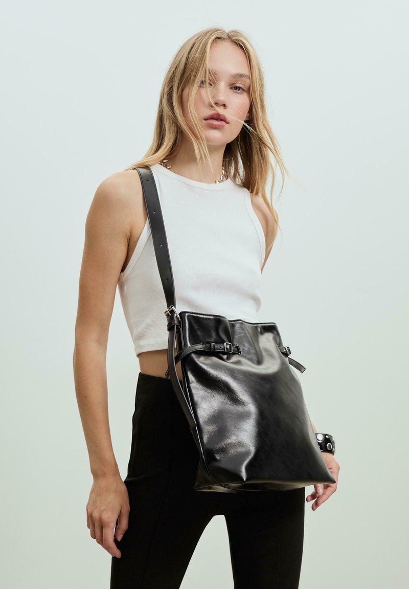 Schwarze Leder-Einkaufs-Tasche mit verstellbarem Riemen, glänzender Oberfläche, gerafftem oberen Teil und zwei seitlichen Schnallen zum Verschließen.