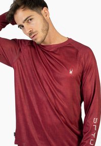 Punainen pitkähihaisen urheilupaidan, joka on valmistettu kevyestä kankaasta, ja siinä on tyylikäs muotoilu, teksturoidut kuviot sekä logo rinnassa ja hihassa.