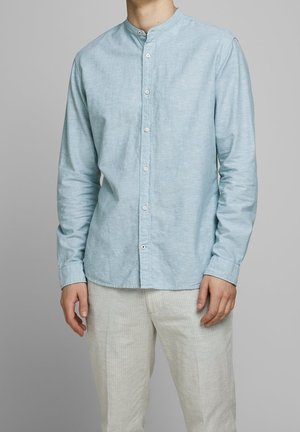 Homme portant une chemise bleu clair à manches longues boutonnée avec un col mao et un pantalon beige clair, debout devant un fond gris.