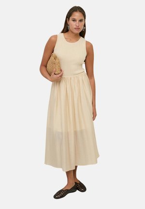 Mujer con vestido midi beige sin mangas sosteniendo un clutch tejido, combinado con bailarinas tejidas negras, de pie sobre un fondo claro liso.