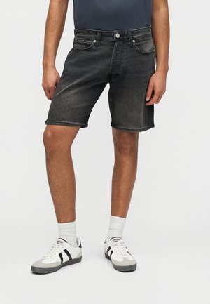 Mand iført sorte denimshorts, hvide sokker og hvide sneakers med sorte striber, stående mod en ensfarvet baggrund.