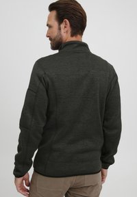 Veste en polaire vert foncé avec un col haut, surface texturée et manches ajustées, présentant des coutures contrastantes le long du dos et des épaules.