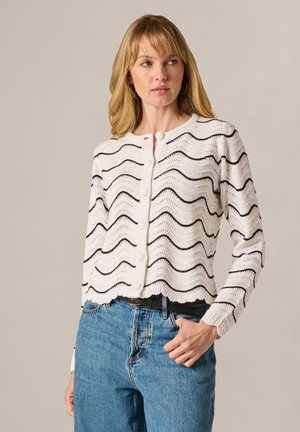 Woman wearing white knit cardigan with black wavy stripes and buttons, paired with blue jeans and a black belt, standing against plain background.