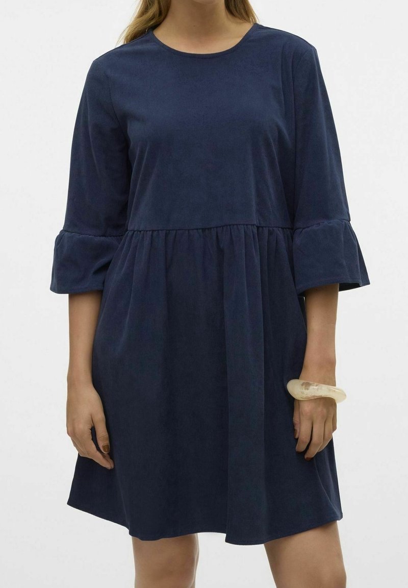 Femme portant une robe en velours côtelé bleu marine avec des manches cloches trois-quarts et une taille froncée, accessoirisée d’un bracelet translucide épais.