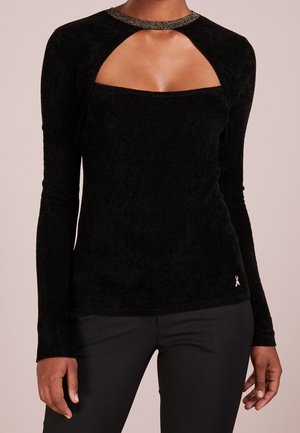 Haut noir à manches longues avec col et découpe pailletés sur la poitrine, porté avec un pantalon noir par une personne debout à la peau brune.