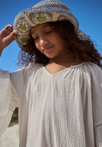Blusa bianca con trama, scollo a V, maniche ampie e cappello all'uncinetto blu, beige e verde su uno sfondo di cielo azzurro limpido.